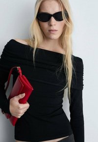Top negru fără umeri, dintr-un material texturat, cu mâneci lungi, asortat cu o geantă roșie de forma rectangulară. Modelul poartă ochelari de soare închiși la culoare.