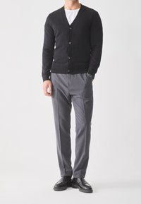 Svart cardigan med V-hals och knappstängning, kombinerad med grå, plisserade byxor. Modellen bär svarta skor mot en neutral bakgrund.