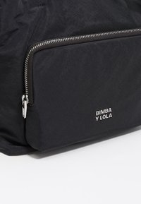 Zbliżenie kieszeni czarnej torby z srebrnym zamkiem błyskawicznym i małym metalowym logo "BIMBA Y LOLA" na tkaninie.