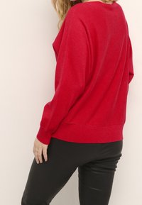 Pull oversize en maille rouge avec épaules tombantes, poignets et ourlet côtelés, associé à un pantalon noir ajusté. Texture douce, design décontracté.