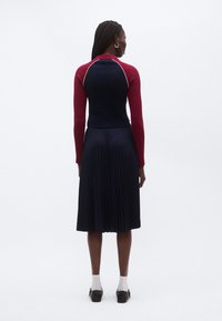 Sukeldumisvormiga ja pikkade punaste varrukatega kudumispusa, millel on tumesinise kehaga ja volditud tumesinine seelik. Omadused: sooniline tekstuur ja kontrastsed õmblused.