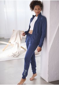 Marineblauwe zip-up jas met hoge kraag en bijpassende joggers, beide van zachte stof. De broek heeft het 'Bench'-script op het onderbeen.