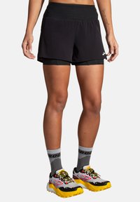 Shorts de sport noirs avec une taille large ; design superposé avec un short intérieur noir. Associés à des chaussettes grises et des chaussures de course multicolores.