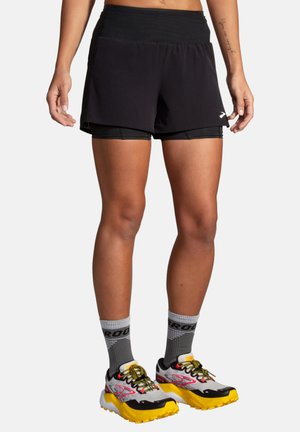 Pantalones cortos deportivos negros con una cinturilla ancha; diseño en capas que incluye un sujetador interior negro. Combinados con calcetines grises y zapatillas de correr multicolores.
