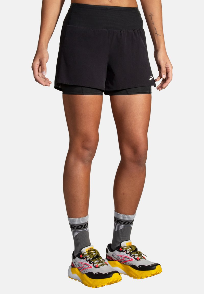Shorts de sport noirs avec une taille large ; design superposé avec un short intérieur noir. Associés à des chaussettes grises et des chaussures de course multicolores.