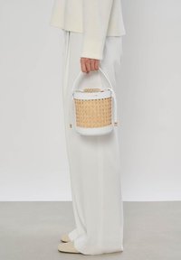 Personne vêtue de blanc tenant un petit sac à main cylindrique en osier et cuir blanc avec une poignée supérieure sur un fond gris uni.