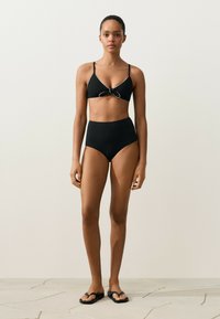 Vrouw die staat en een zwarte bikini draagt met een voorgeknoopte top en een broekje met hoge taille en zwarte teenslippers tegen een lichte muur.