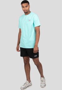 Camiseta de algodón turquesa con cuello redondo y pequeño logo en el pecho, combinada con pantalones cortos negros que tienen texto en blanco. Zapatillas grises.