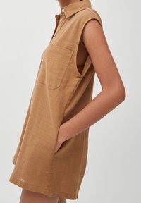 Robe chemise marron sans manches en tissu texturé, avec un col chemise, une poche poitrine et des poches latérales.
