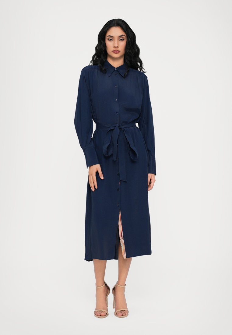 Robe bleu marine à boutons avec des manches longues, ceinture à la taille et longueur mi-mollet. Présente une fente avant révélant une doublure à motifs en dessous.