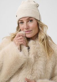 Manteau en fausse fourrure beige clair avec de longues fibres douces et des manches larges ; un bonnet en tricot crème avec un motif texturé subtil.