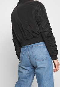 Černá bomber bunda se štíhlým designem, přiléhavými manžetami a lehkým leskem. V kombinaci s vysokým světlým modrým denimem.