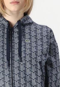 Námornícky zipsový mikina s geometrickým bielym vzorom, kapucňou so sťahovacou šnúrkou a malým zeleným logom na hrudi. Hladká textúra látky.