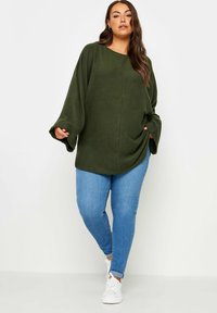 Olivengrön, oversized tröja med breda ärmar, mjuk textur, kombinerad med ljusblå jeans och vita sneakers.