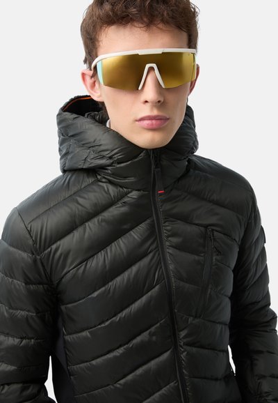 Schwarze Daunenjacke mit gestepptem Muster und frontalem Reißverschluss. Verfügt über eine Kapuze und eine Brusttasche. Die Sonnenbrille hat einen weißen Rahmen und gelbe Gläser.
