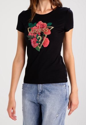 Kobieta w czarnej koszulce Guess z brokatowym logo i różowym nadrukiem róż, w połączeniu z jasnoniebieskimi, postrzępionymi jeansami.
