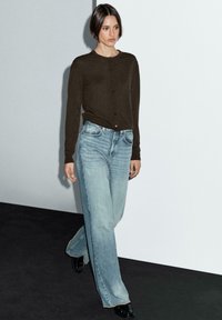 Woman walking indoors wearing a dark brown buttoned cardigan, light blue wide-leg jeans, and black shoes against a white wall background.