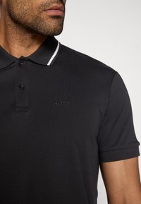 Černé polo tričko s třemi knoflíky, límcem s bílými pruhy a vyšitým logem "BOSS" na hrudi. Hladká textura tkaniny.