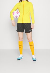 Nike keltainen vetoketjullinen takki, mustat shortsit, keltaiset polvisukat ruskeilla raidoilla ja siniset jalkapallokengät. Pitelee vaaleanpunaista ja valkoista jalkapalloa.
