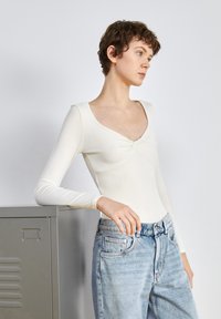 Body a maniche lunghe di colore off-white, con scollatura profonda e dettaglio annodato davanti, abbinato a jeans in denim azzurro chiaro a vita alta.
