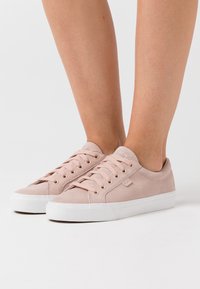 Světlé růžové semišové tenisky s bílou gumovou podešví; mají šest průchodek a ploché tkaničky. Zaoblená špička a minimalistické značení na boku.