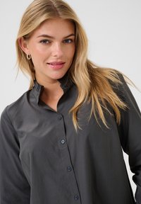Donkergrijze blouse met een staande kraag, knoopsluiting aan de voorkant en lange mouwen. Gladde stof met een subtiele glans en eenvoudige knoopaccenten.