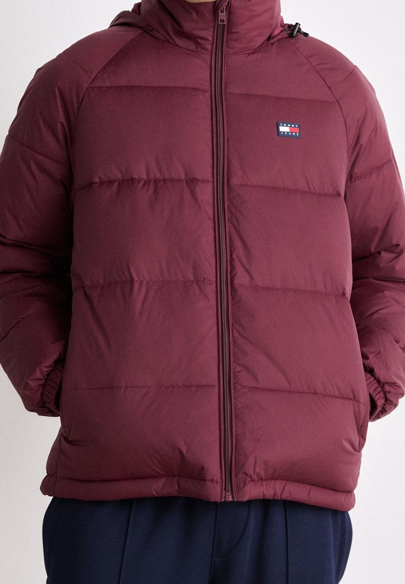 Giacca imbottita bordeaux con zip frontale, design trapuntato e patch con logo sul lato sinistro del petto. Presenta un cappuccio e polsini elasticizzati.