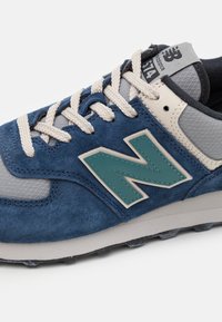 Baskets en daim bleu marine avec des panneaux en maille grise, des lacets crème et un logo turquoise. Les accents comprennent une étiquette noire sur la languette.