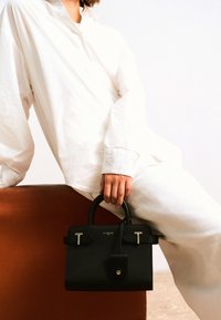 Sac à main noir texturé avec des poignées arrondies et des accents métalliques. Détail de fermoir notable ; exposé sur un fond neutre avec un léger éclat.