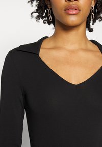 T-shirt preto de mangas compridas com um decote em V e colarinho. Tecido liso, design ajustado. Brincos de corrente dourada com um padrão retangular.