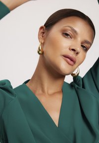 Zielony, szmaragdowy top z falbaniastymi rękawami. Złote kolczyki w kształcie kropli. Gładki materiał o delikatnym połysku, dopasowany krój podkreślający dekolt.