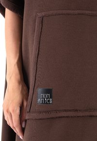 Brązowy, oversize'owy element odzieży z dużą kieszenią na przodzie oraz czarną metką z białym tekstem identyfikującym markę. Miękka tekstura materiału.