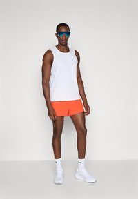 Weißes ärmelloses Sportoberteil, orangefarbene Shorts, blaue Sonnenbrille, weiße Sneakers und weiße Socken. Glattes Material, körperbetontes Design, moderner Look.