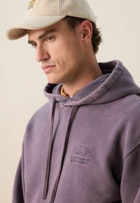 Jonge man met een beige pet en een paarse hoodie met geborduurde tekst, die naar beneden kijkt tegen een effen lichte achtergrond.