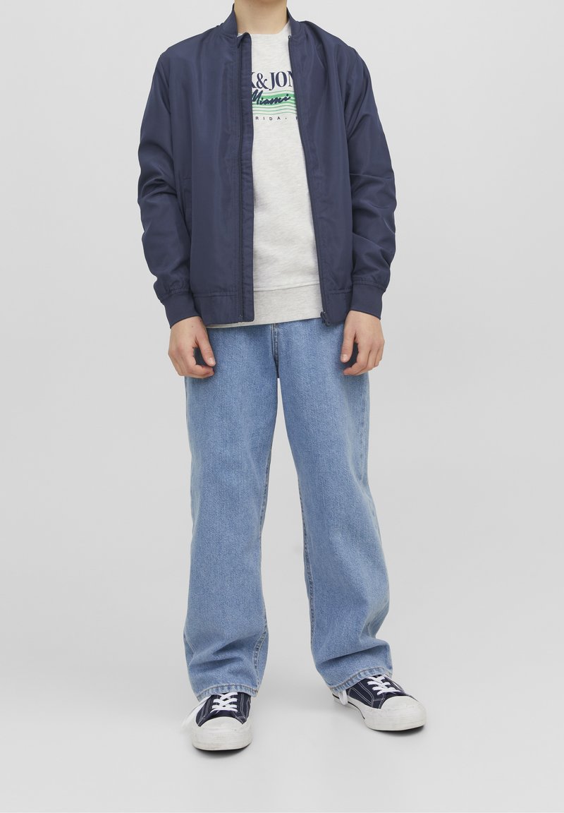 Granatowa kurtka bombowa, szary sweter z napisem, jasnoniebieskie szerokie dżinsy i czarno-białe trampki ze sznurówkami. Gładki materiał, luźny krój.