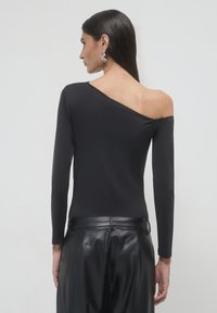 Donna con lunghi capelli scuri che indossa un top lungo a maniche lunghe asimmetrico con spalla scoperta di colore nero e pantaloni in pelle nera, voltata di spalle su uno sfondo semplice.