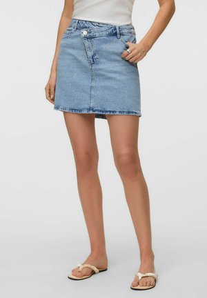Femme portant une mini-jupe en jean asymétrique bleu clair, un débardeur blanc et des sandales beiges avec des lanières décoratives.