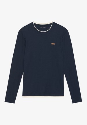 Garā piedurkņu tumši zila ribota krekls ar baltu apkakli, kuram ir neliela brūna birka uz krūtīm. Mīksta tekstūra un brīva piegriezuma.