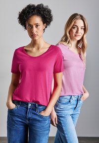 Dos mujeres visten camisetas de manga corta en tonos de rosa, una rosa brillante y la otra rosa claro, combinadas con jeans azules.