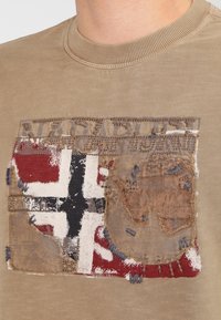 T-shirt a maniche corte color khaki con un patch testurizzato che presenta un design di bandiera nei colori nero, bianco, rosso e marrone, arricchito da dettagli di cucitura visibili.