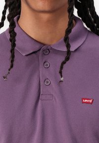 Tricou polo mov cu guler, patru butoane și un mic logo Levi's roșu pe piept. Materialul pare texturat și țesut.
