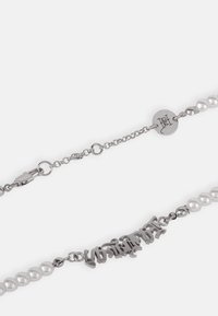 Colar de corrente prateada com pérolas brancas e um centro com inscrição metálica. Inclui um fecho e uma pequena etiqueta com o logótipo.