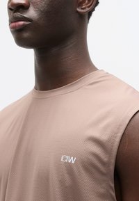 Καφέ αμάνικο αθλητικό μπλουζάκι από υφασμάτινη υφή, με ένα μικρό λευκό λογότυπο "ICIW" στην αριστερή πλευρά του στήθους.