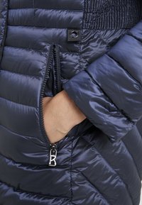 Laivastonsininen toppatakki tikatulla pinnalla, jossa on vetoketjullinen tasku, pystykaulus ja hopeiset metalliosat. Sileä, kiiltävä materiaali.