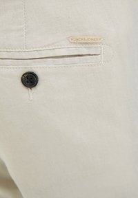 Gros plan sur un pantalon en tissu beige avec une poche arrière boutonnée et une petite étiquette "JACK & JONES" au-dessus de la poche.