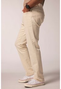 Beige Baumwolljeans mit geradem Schnitt, ausgestattet mit Vordertaschen und subtilen Stickdetails, kombiniert mit weißen Sneakers.
