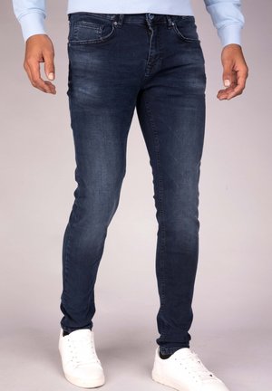 Homme portant un jean slim bleu foncé et des baskets blanches, debout avec les bras détendus le long du corps devant un fond uni.
