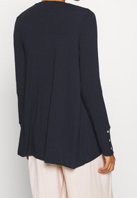 Granatowy top z długim rękawem wykonany z miękkiej tkaniny, z zaokrąglonym dołem i mankietami zapinanymi na guziki, noszony na jasnych spodniach.