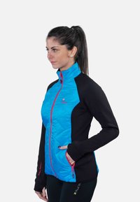 Femme portant une veste zippée bleu et noire avec des accents rouges, debout de profil avec une main dans la poche, sur un fond uni.