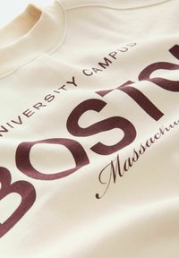 Krämfärgad sweatshirt med stora vinröda texter: "Boston" och "University Campus, Massachusetts." Ribbad halsringning och mjukt material.